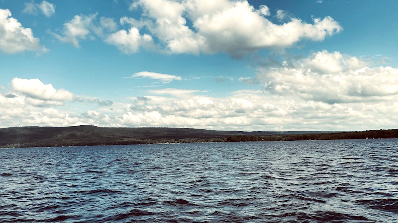 Dark blue Great Sacandaga Lake with light waves looking toward the shoreline with low mountains under a bright blue sky